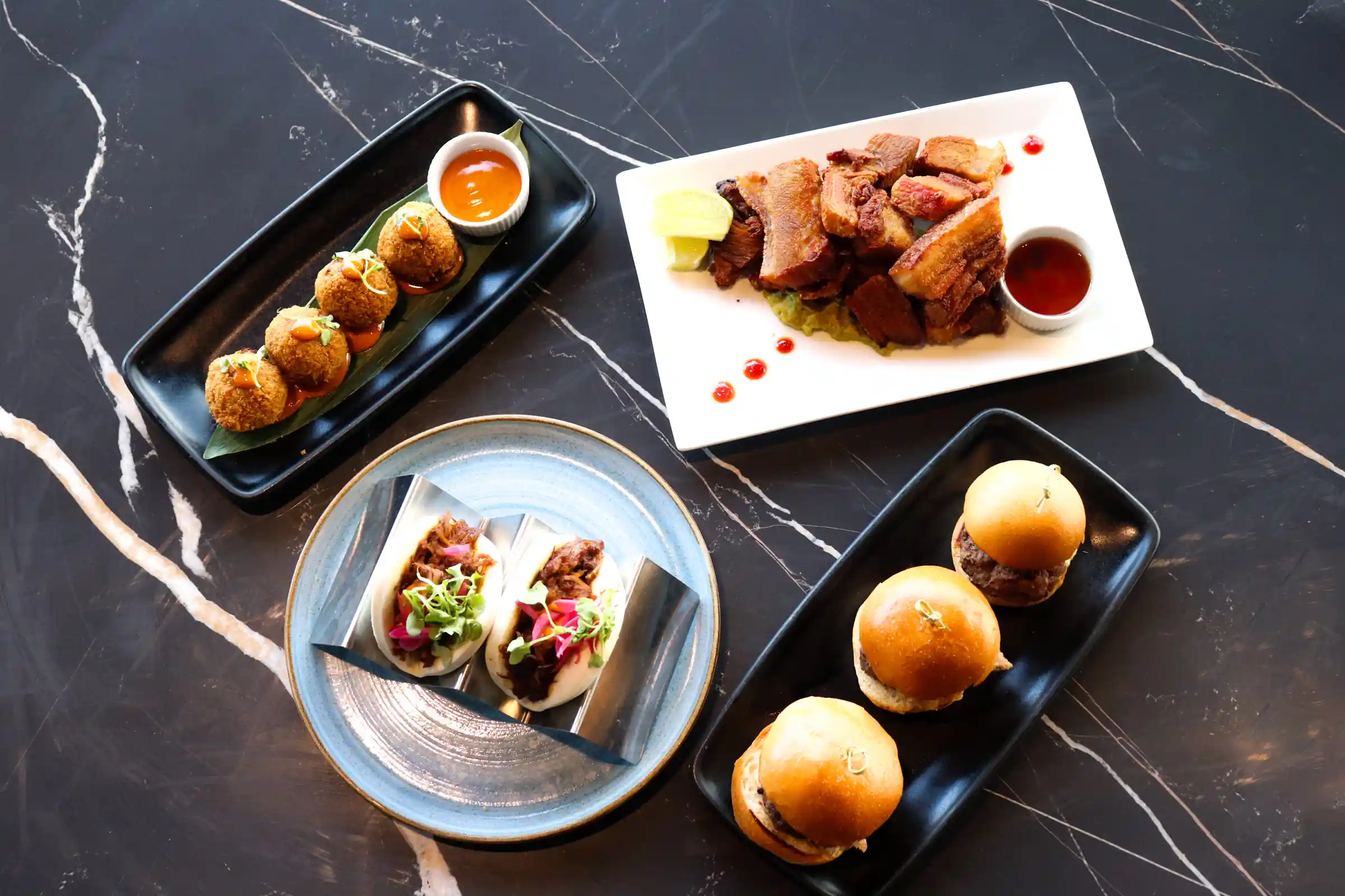 Una variedad de platos gourmet, incluyendo cerdo crujiente, tacos sabrosos y mini hamburguesas, dispuestos artísticamente en una sofisticada mesa de mármol