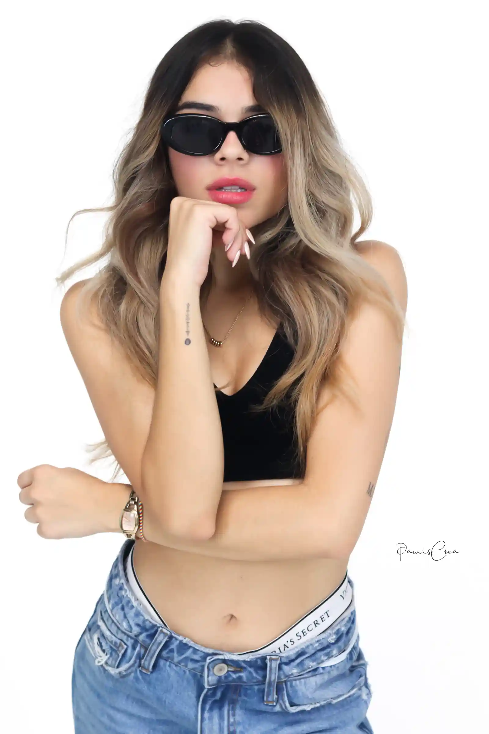 Un retrato de una mujer en una camiseta negra y jeans posa con confianza con cabello ondulado y gafas de sol contra un fondo limpio